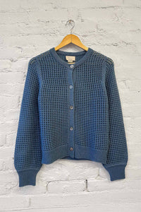 Soft, blue textured cashmere cardigan on a hanger against a white brick wall.