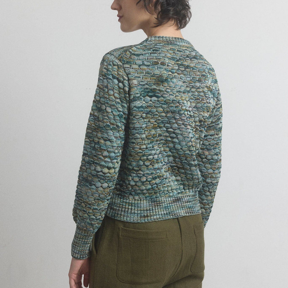 Person wearing a patterned green sweater and olive pants against a plain background