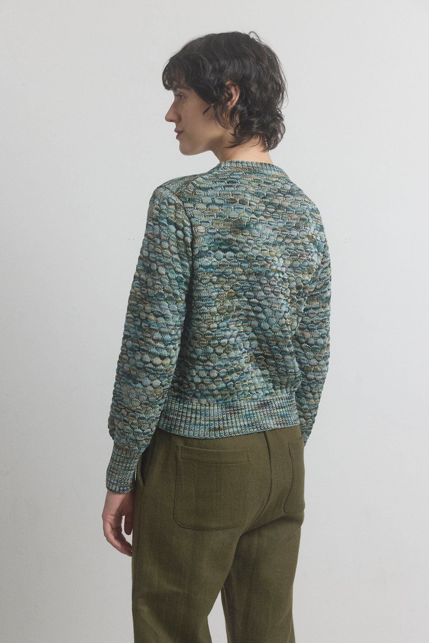 Person wearing a patterned green sweater and olive pants against a plain background
