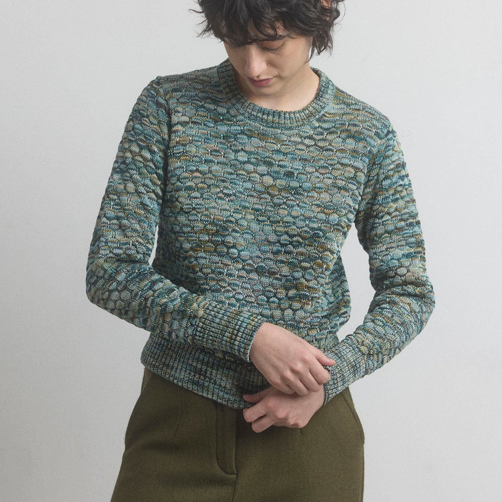 Woman wearing a textured green longn sleeve sweater and olive pants against a plain background