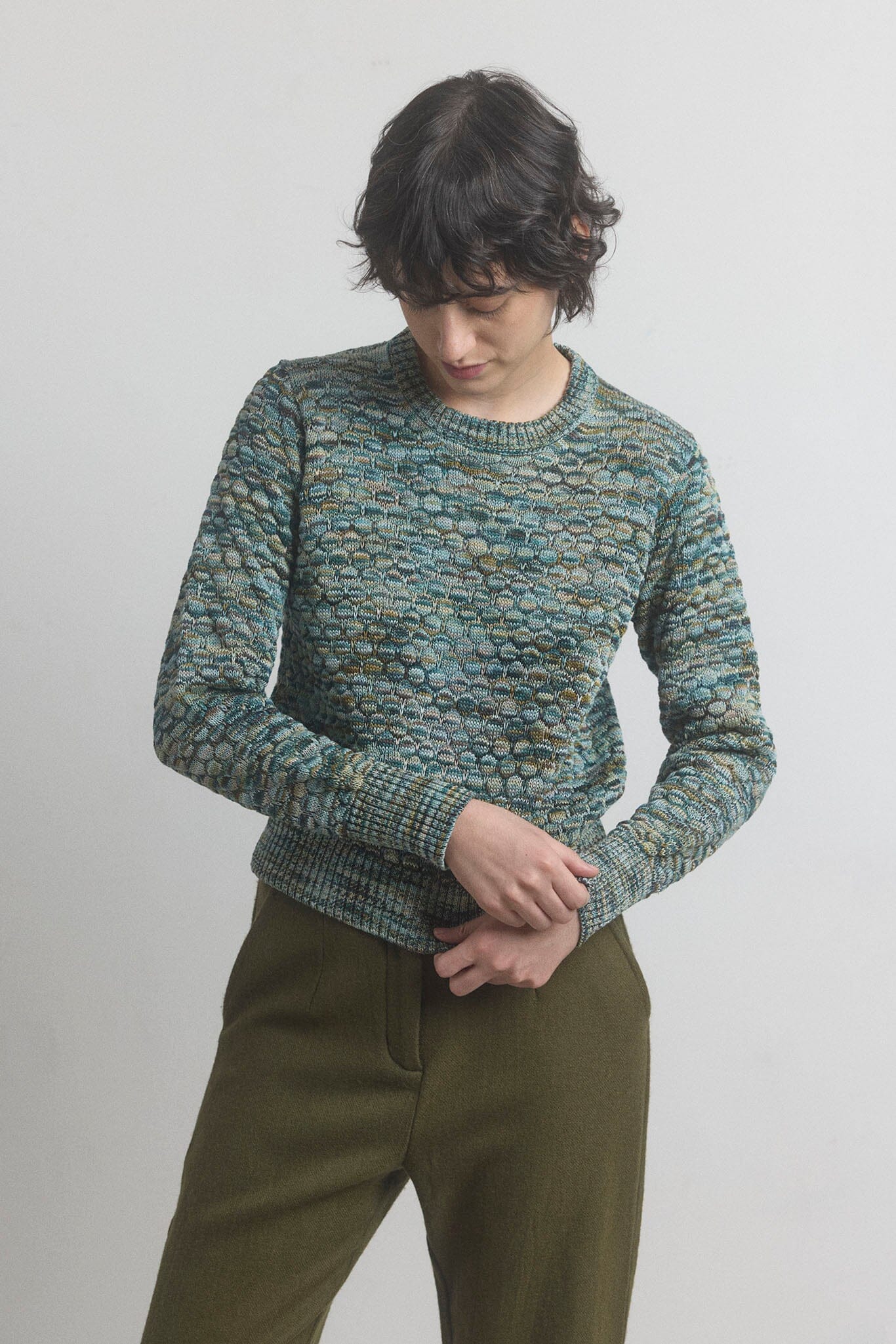 Woman wearing a textured green longn sleeve sweater and olive pants against a plain background