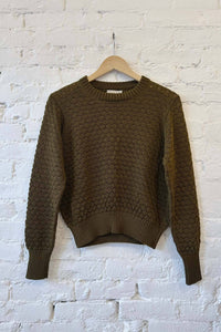 Olive knitted cotton sweater on a wooden hanger against a white brick wall.