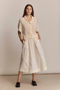 Woman wearing a cream-colored cardigan and ivory skirt against a plain background