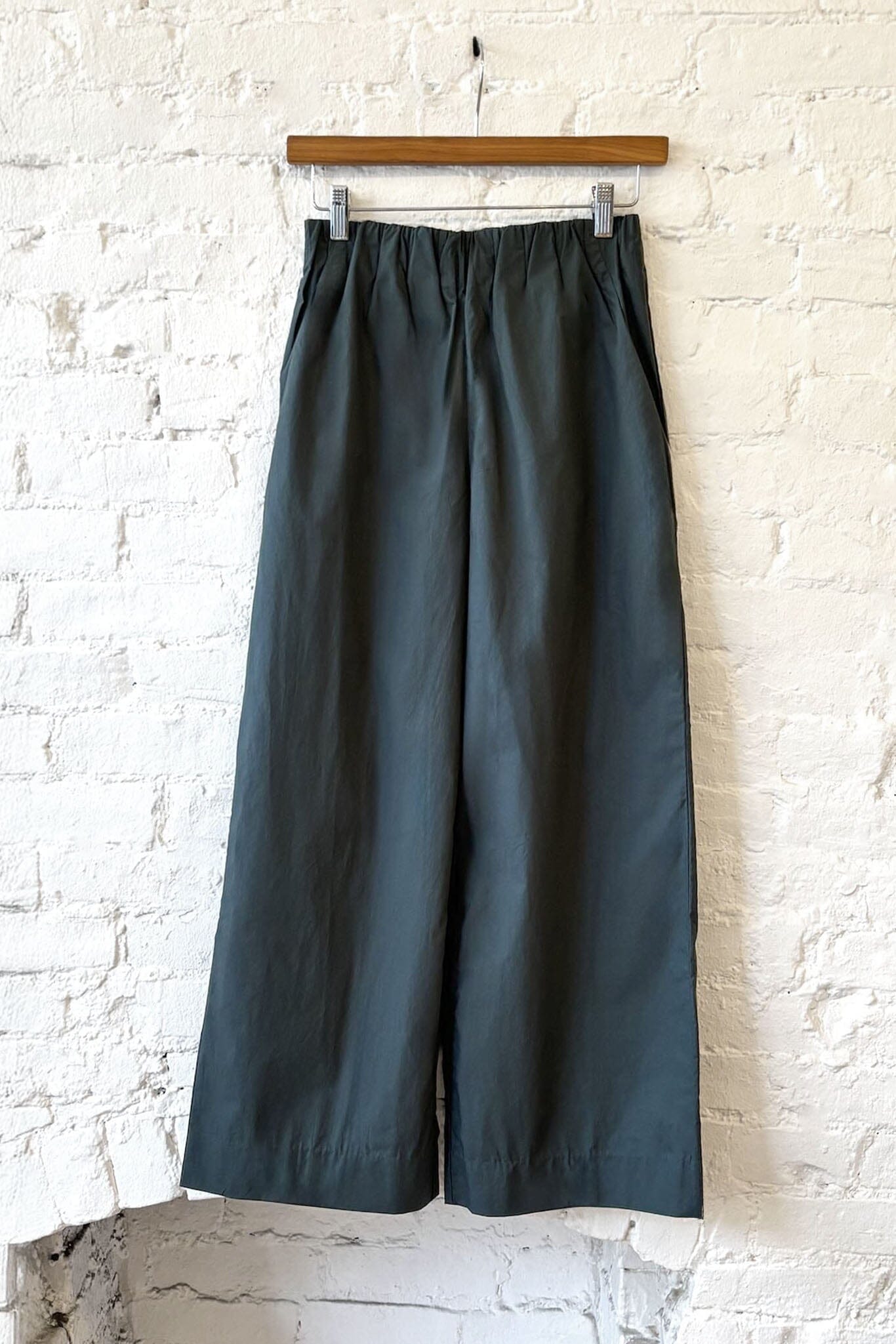 Dark green wide-leg pants hanging on a wooden hanger against a white brick wall.