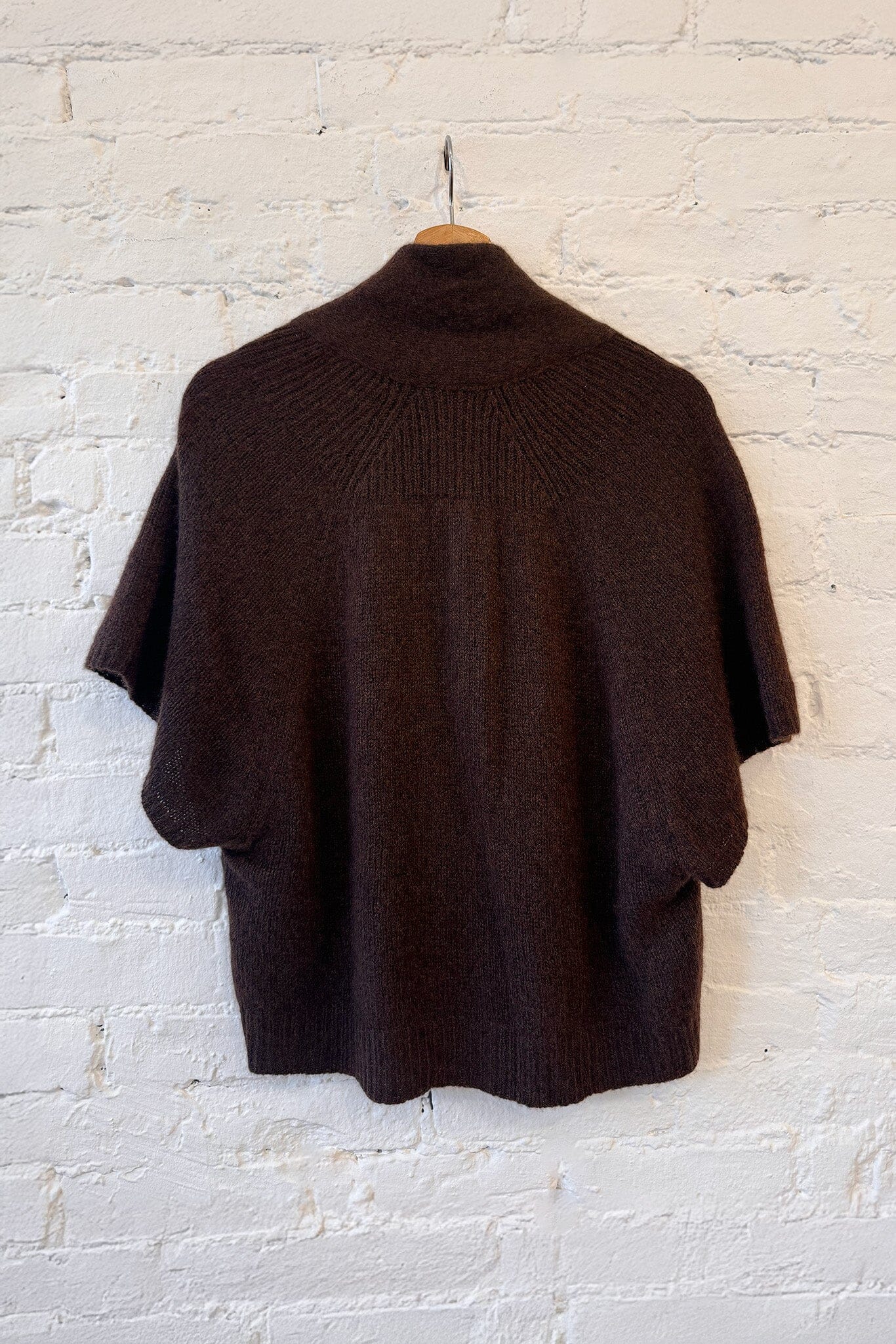 The back of a brown short-sleeve cashmere sweater with mother of pearl buttons on a hanger against a white brick wall.