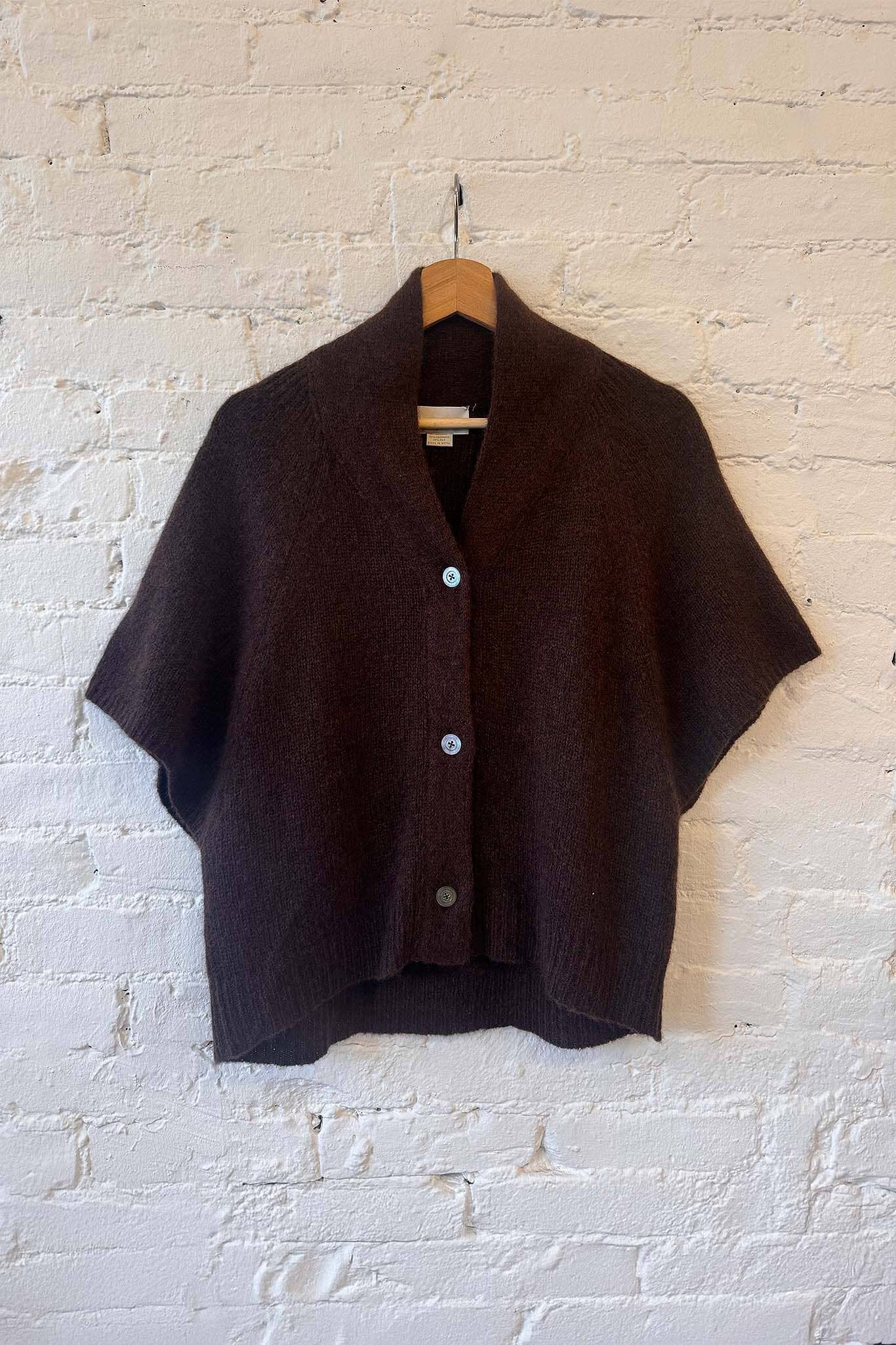 Brown short-sleeve cashmere sweater with mother of pearl buttons on a hanger against a white brick wall.