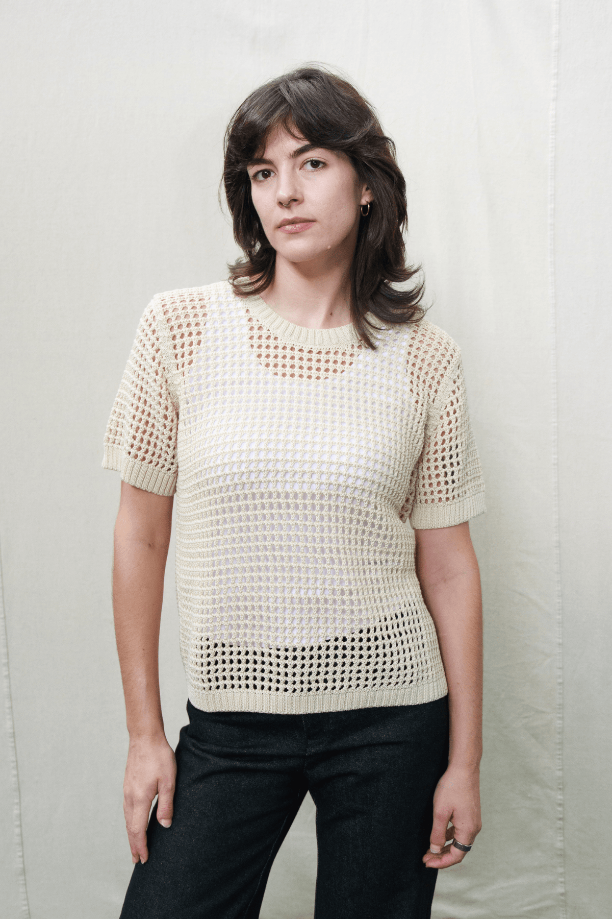 A woman wearing a cream-colored short-sleeve knit top with an open, textured grid pattern and black pants, standing against a light beige fabric backdrop.