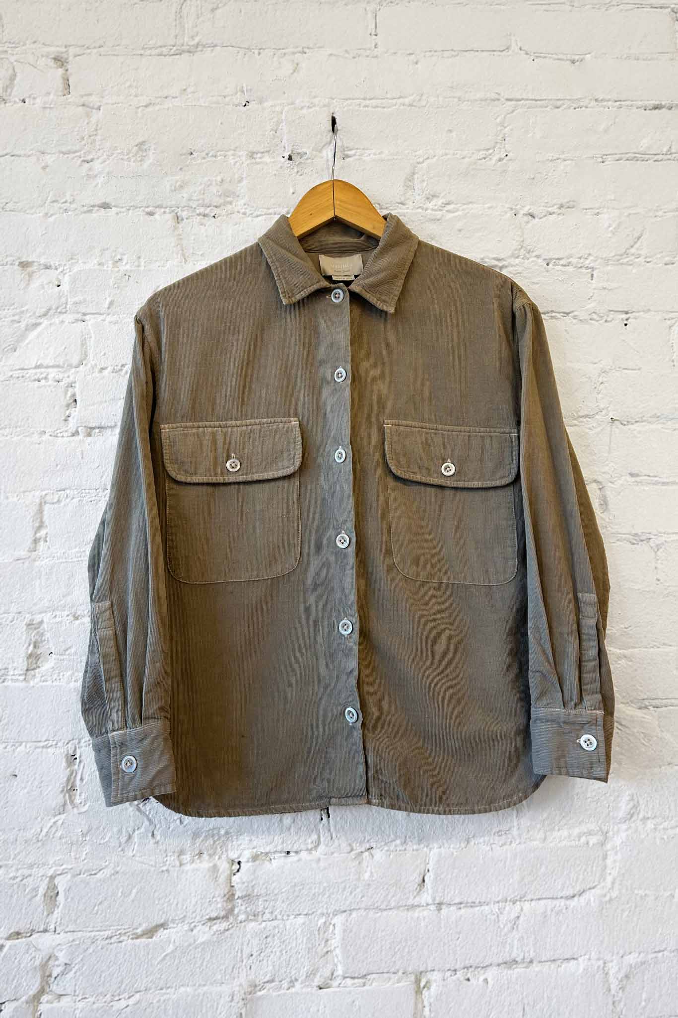 Sage green long sleeve shirt with two front pockets on a wooden hanger against a white brick wall.