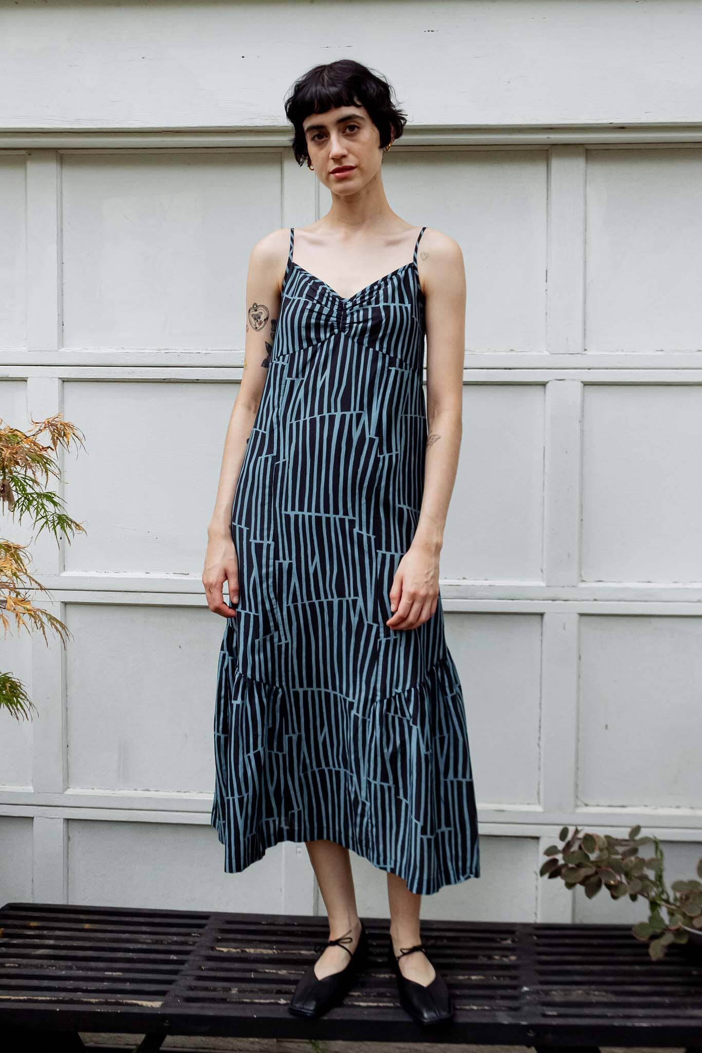 A woman wearing a sleeveless midi dress with thin straps and a vertical blue, black, and white stripe pattern, standing against a white paneled wall.