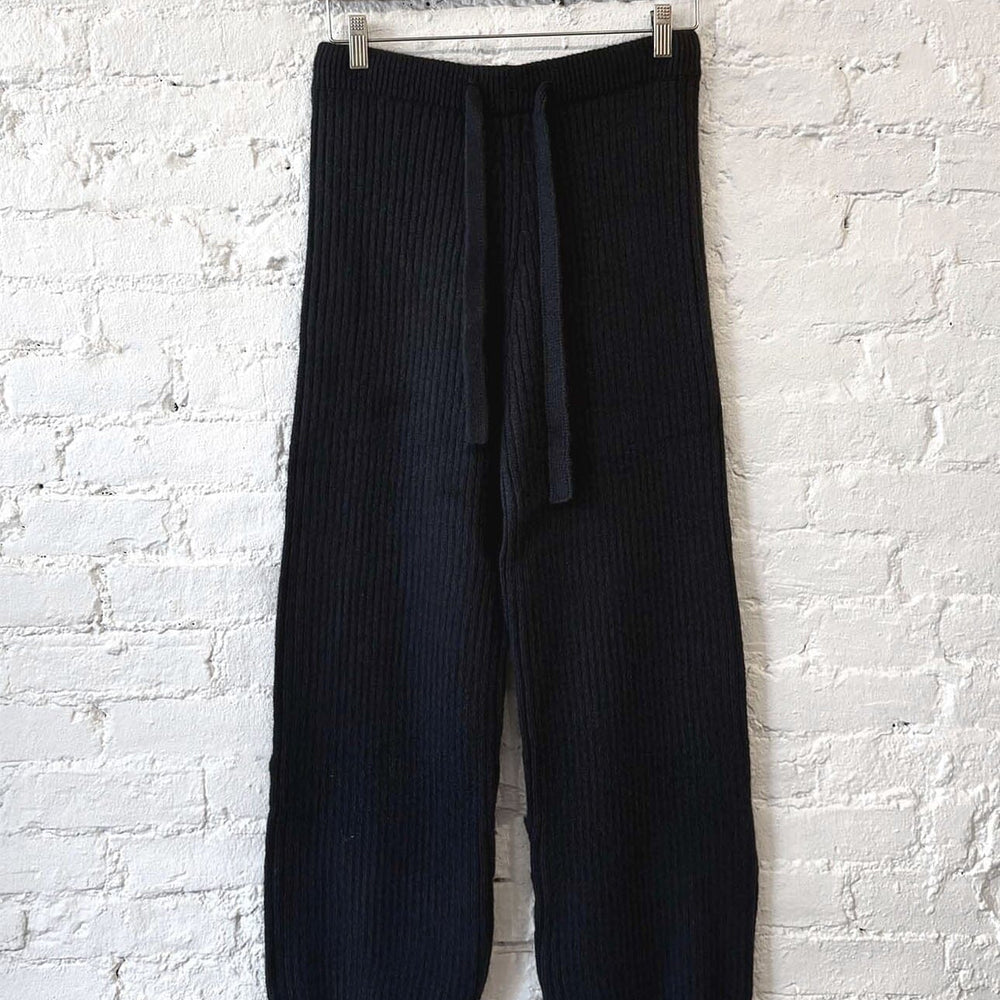 Black ribbed knit pants hanging on a wooden hanger against a white brick wall.