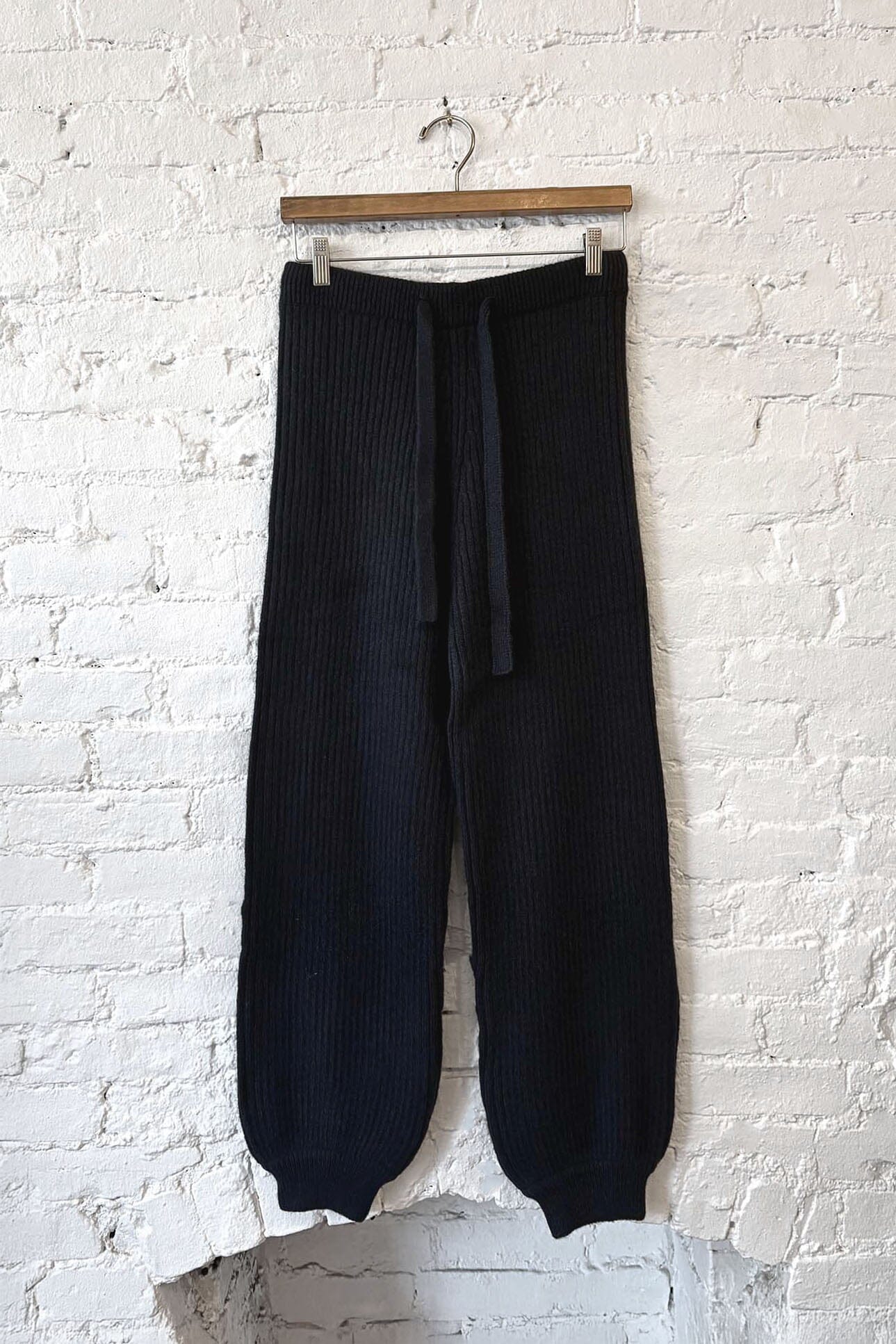 Black ribbed knit pants hanging on a wooden hanger against a white brick wall.