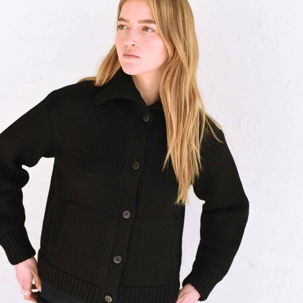 A woman wearing a black textural funnelneck cardigan with natural horn buttons and front welt pockets.
