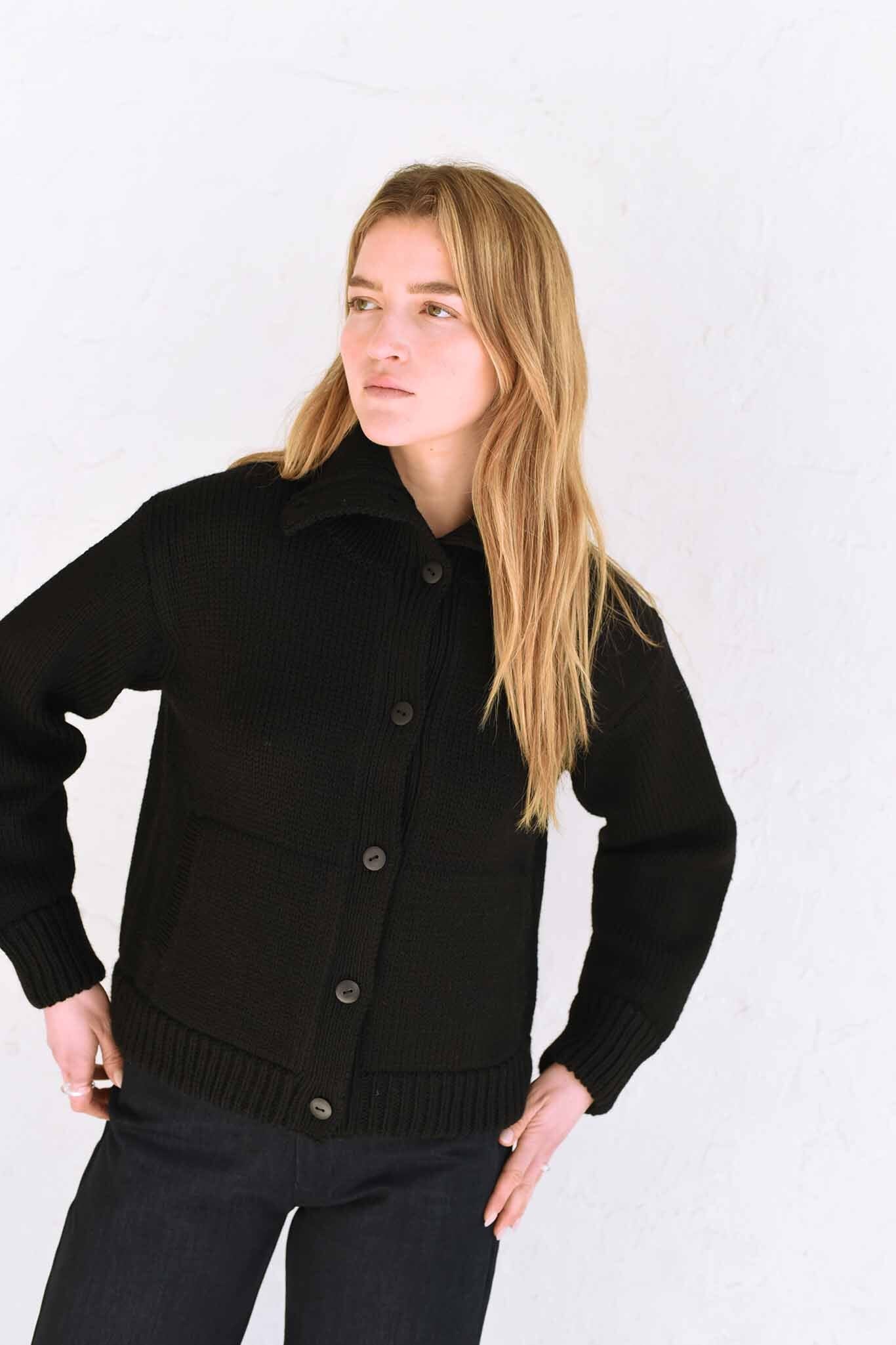 A woman wearing a black textural funnelneck cardigan with natural horn buttons and front welt pockets.
