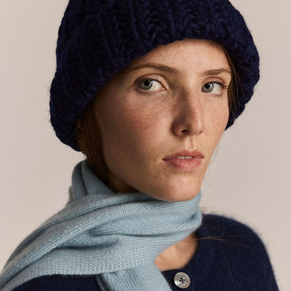 Woman wearing a navy knit beanie and light blue scarf against a beige background