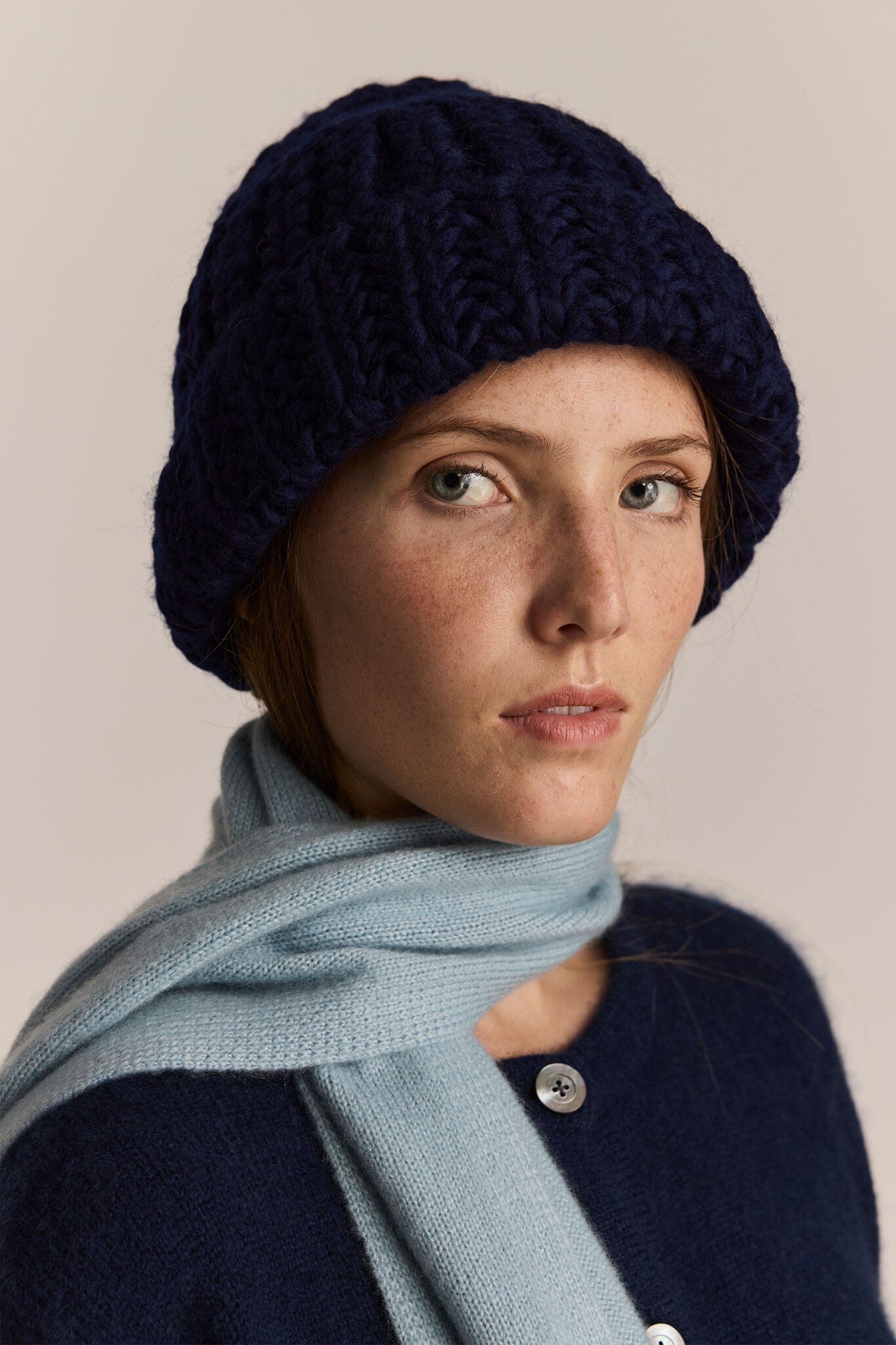 Woman wearing a navy knit beanie and light blue scarf against a beige background