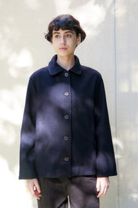 A model wearing a dark navy blue button-up coat with a wide collar, photographed against a light gray wall with soft leaf shadows.