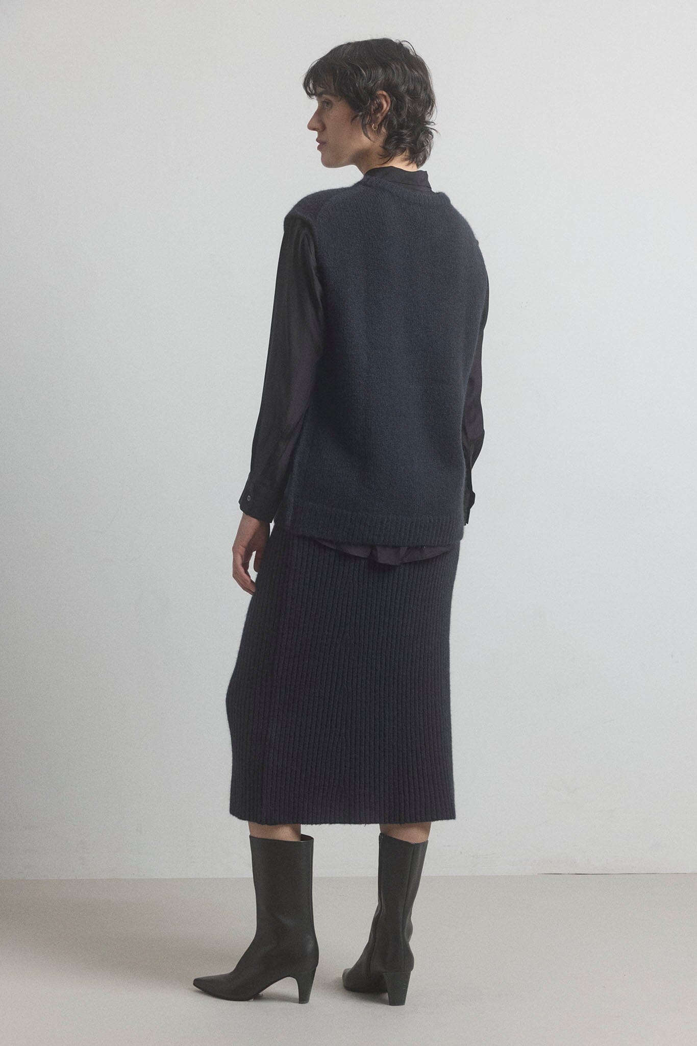 Woman facing the back wearing a black merino vest and skirt on a white background