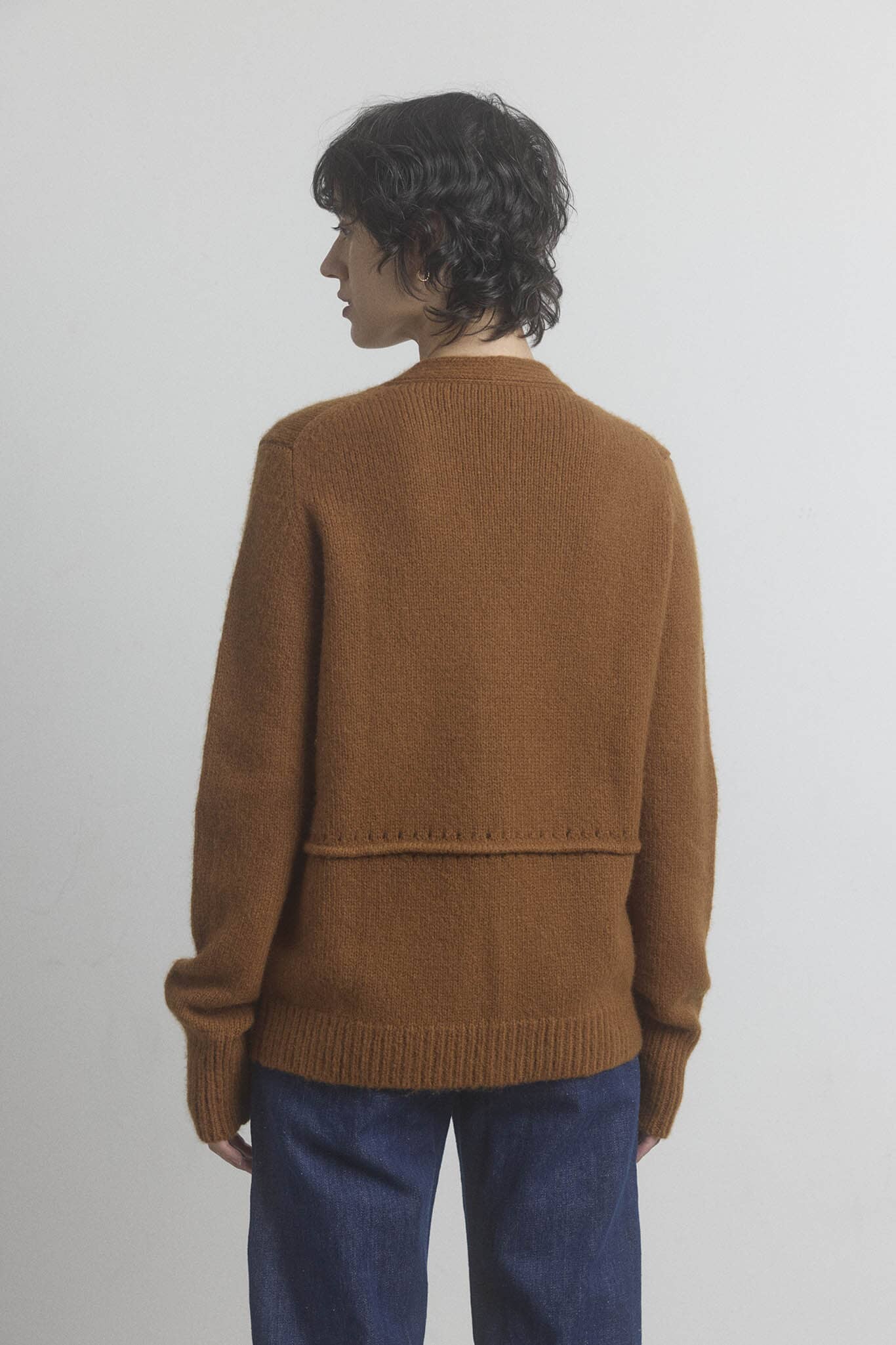Woman wearing a brown cashmere and wool sweater and blue jeans against a plain background.