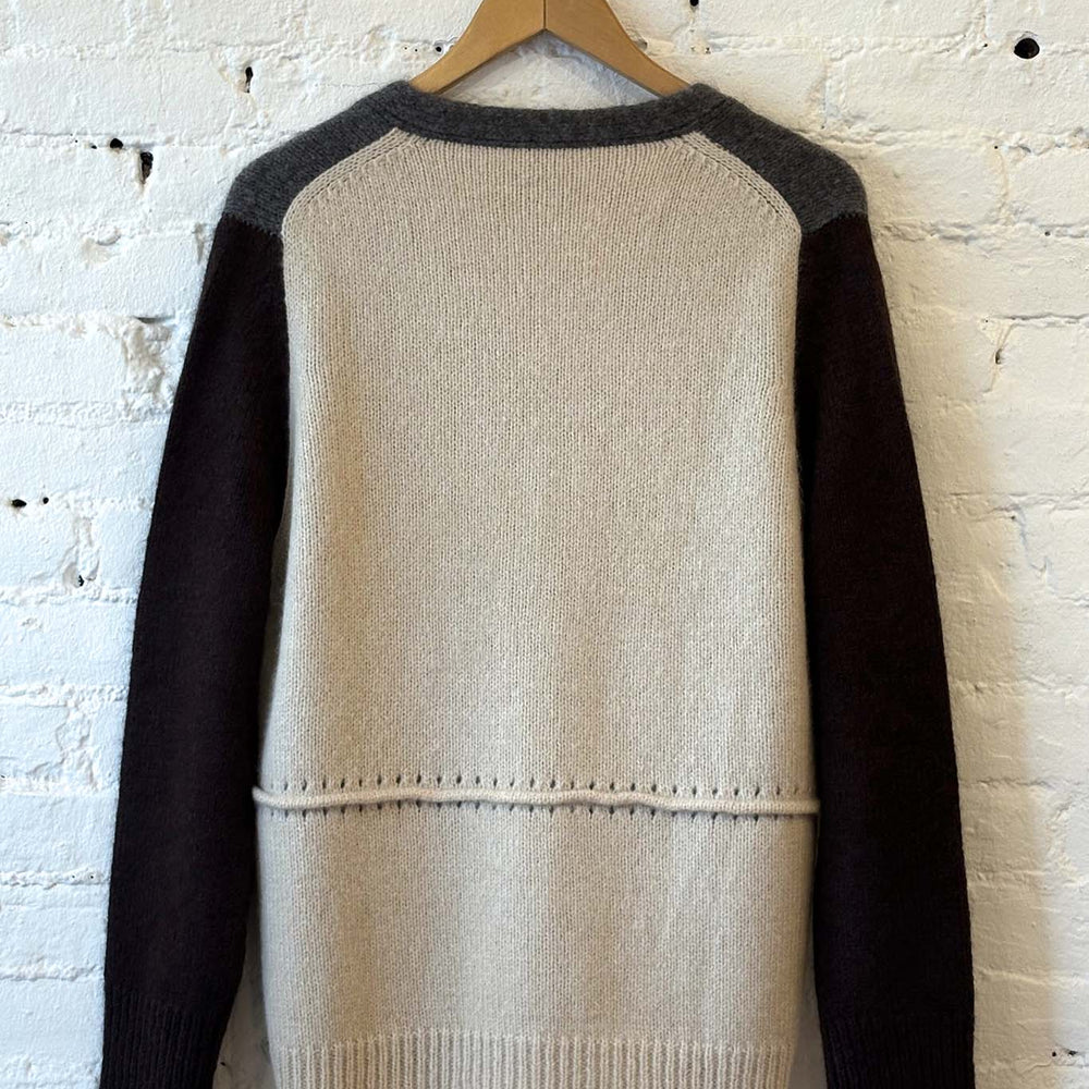 
                  
                    Beige, grey sweater with black sleeves and beige body on a wooden hanger against a white brick wall.
                  
                