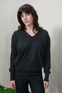 A woman with shoulder-length brown hair wearing a dark charcoal gray V-neck knit sweater and dark jeans, standing against a light beige fabric backdrop.