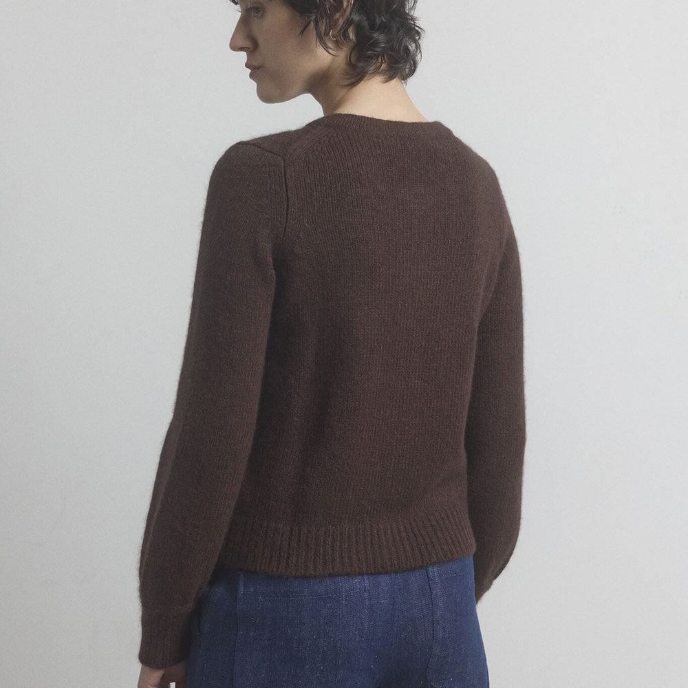Woman facing the back wearing a brown cashmere sweater and blue pants against a plain background
