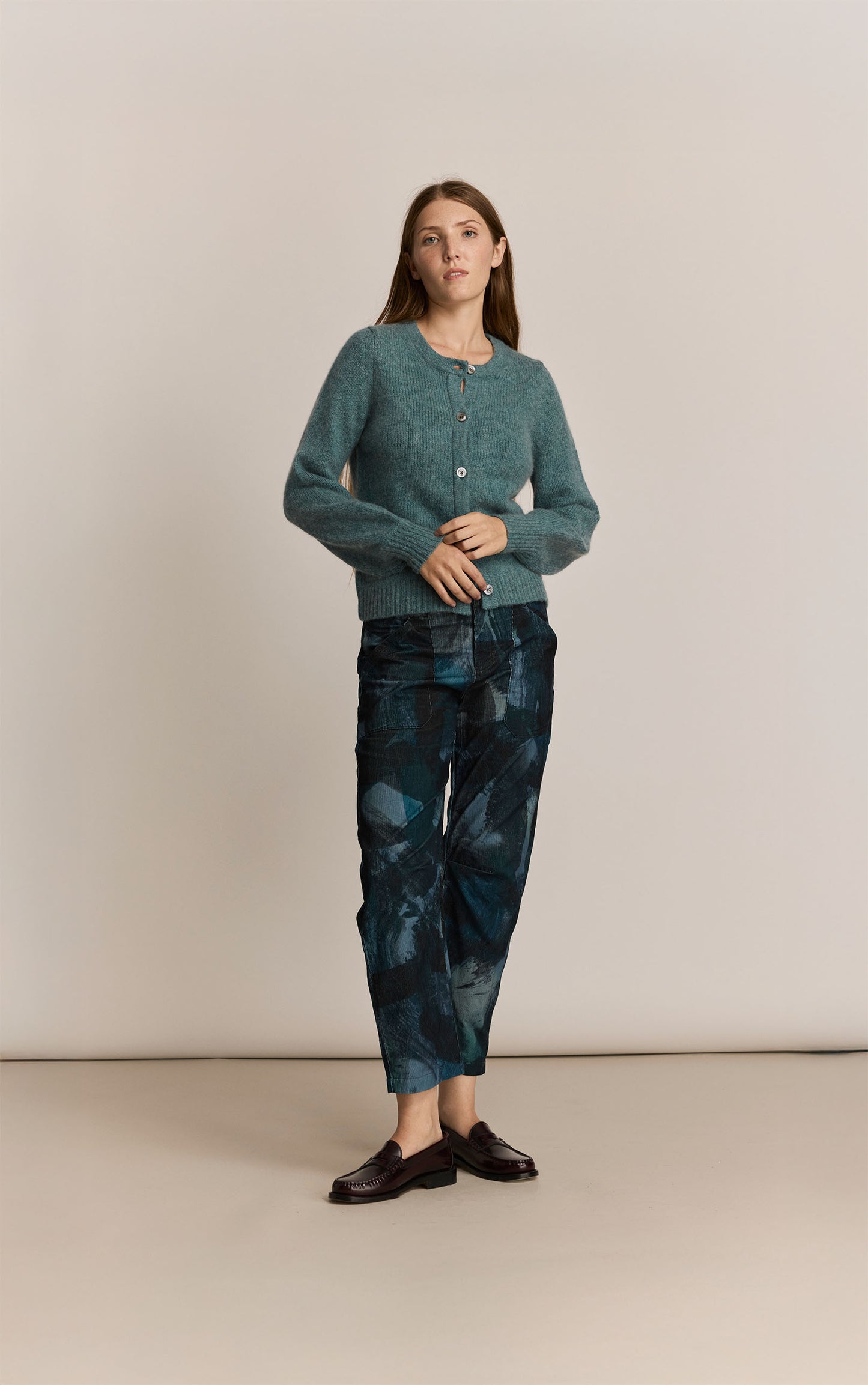 Woman wearing a green knitted cardigan and patterned pants against a beige background