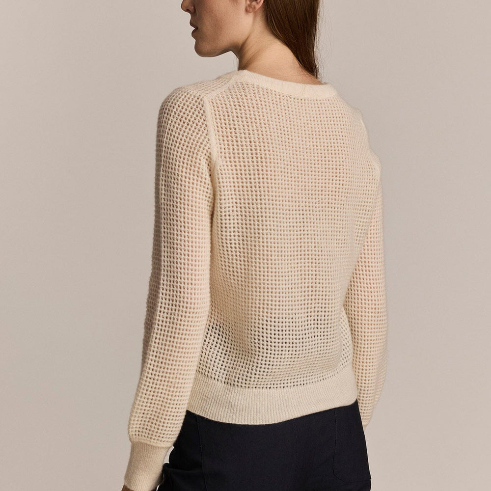 A woman wears an ivory colored, long sleeve mesh sweater against a grey background.