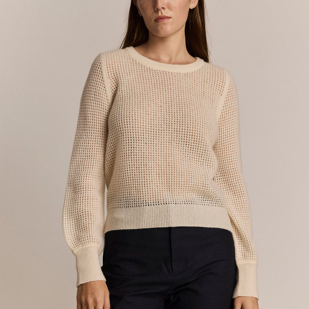 A woman wears an ivory colored, long sleeve mesh sweater against a grey background.