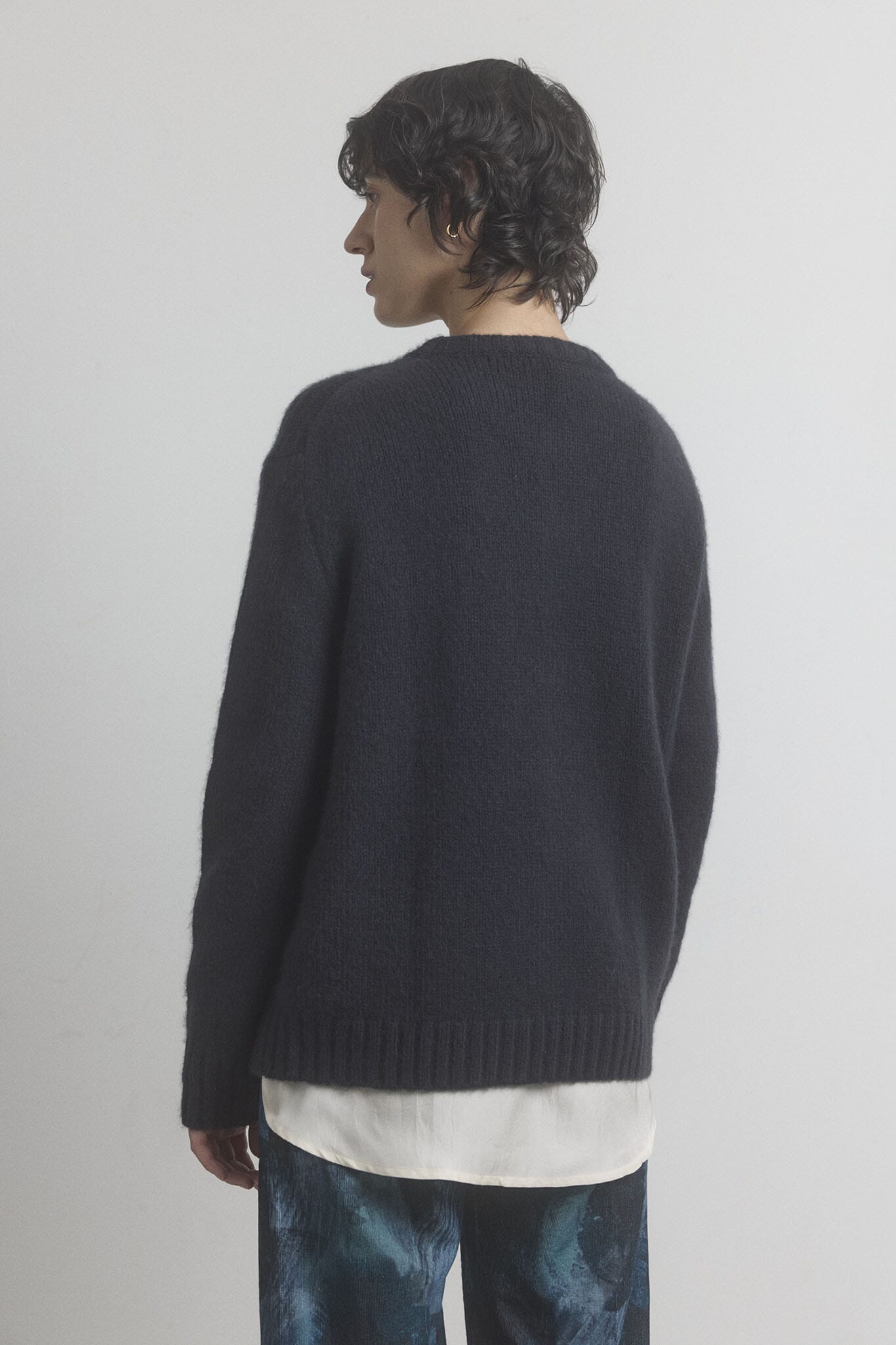 Woman facing the back wearing a merino wool sweater over a white shirt with a plain background