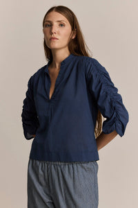 Woman wearing a blue blouse with long, gathered detailing on the sleeves against a beige background