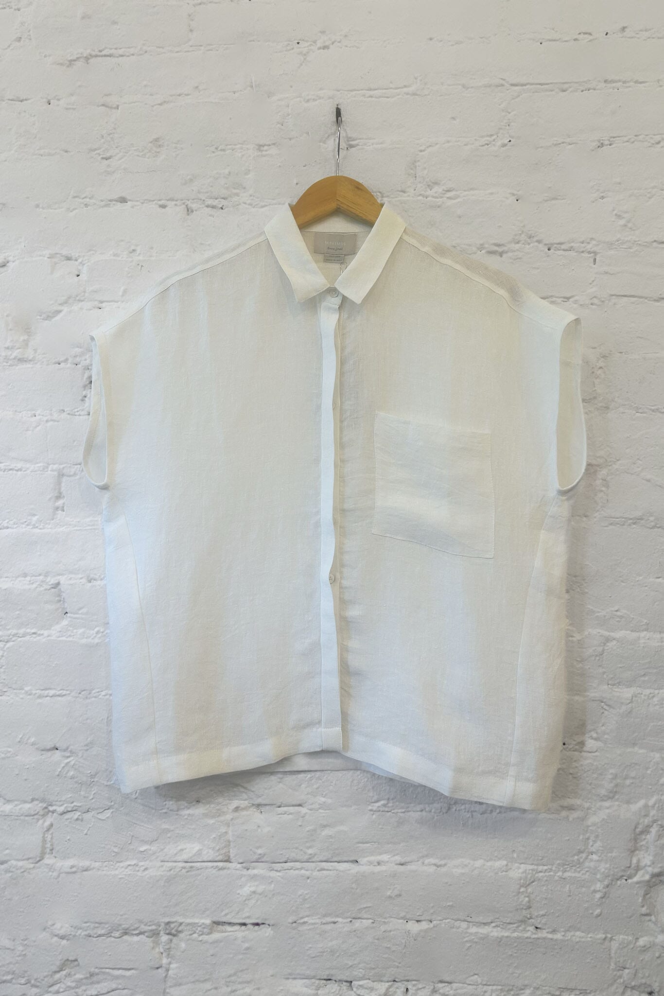 Lightweight, white short sleeve blouse on a wooden hanger against a white brick wall.
