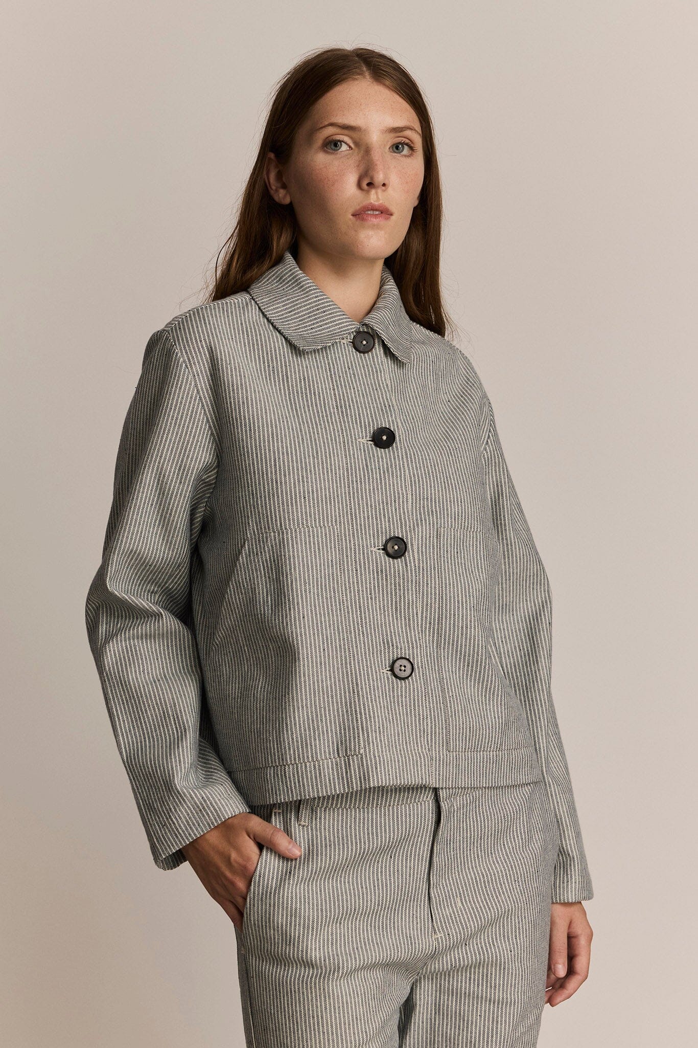 Woman wearing a cropped grey striped chore jacket against a beige background