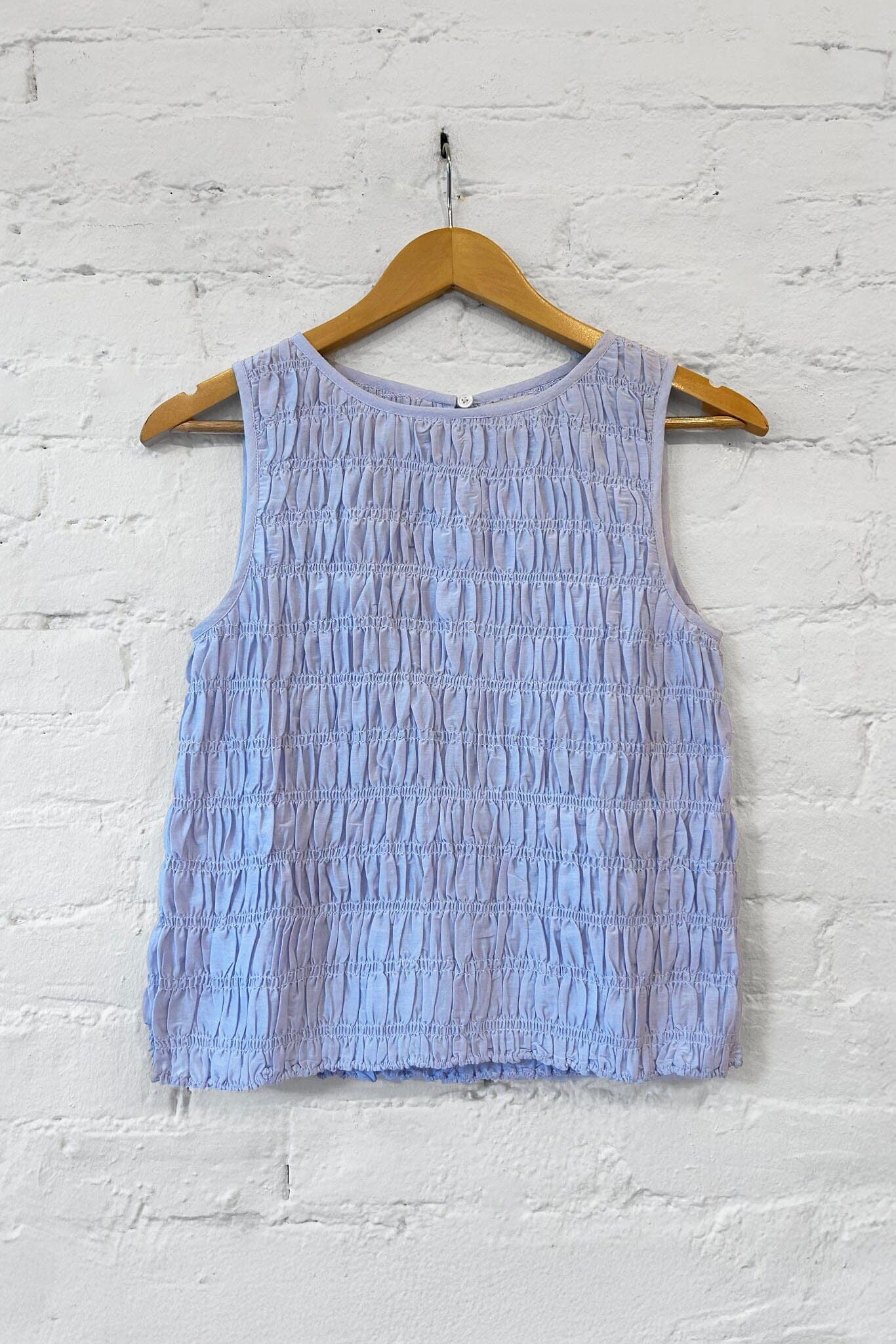 Blue sleeveless top on a wooden hanger against a white brick wall.