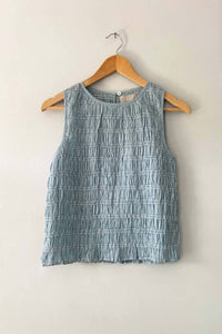 A sleeveless smocked top in a dusty light blue-gray color, hanging on a wooden hanger against a plain off-white wall.