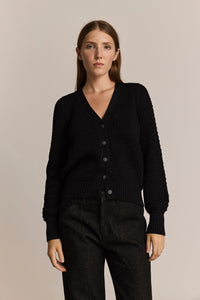 Woman wearing a black cardigan and navy denim pants against a beige background