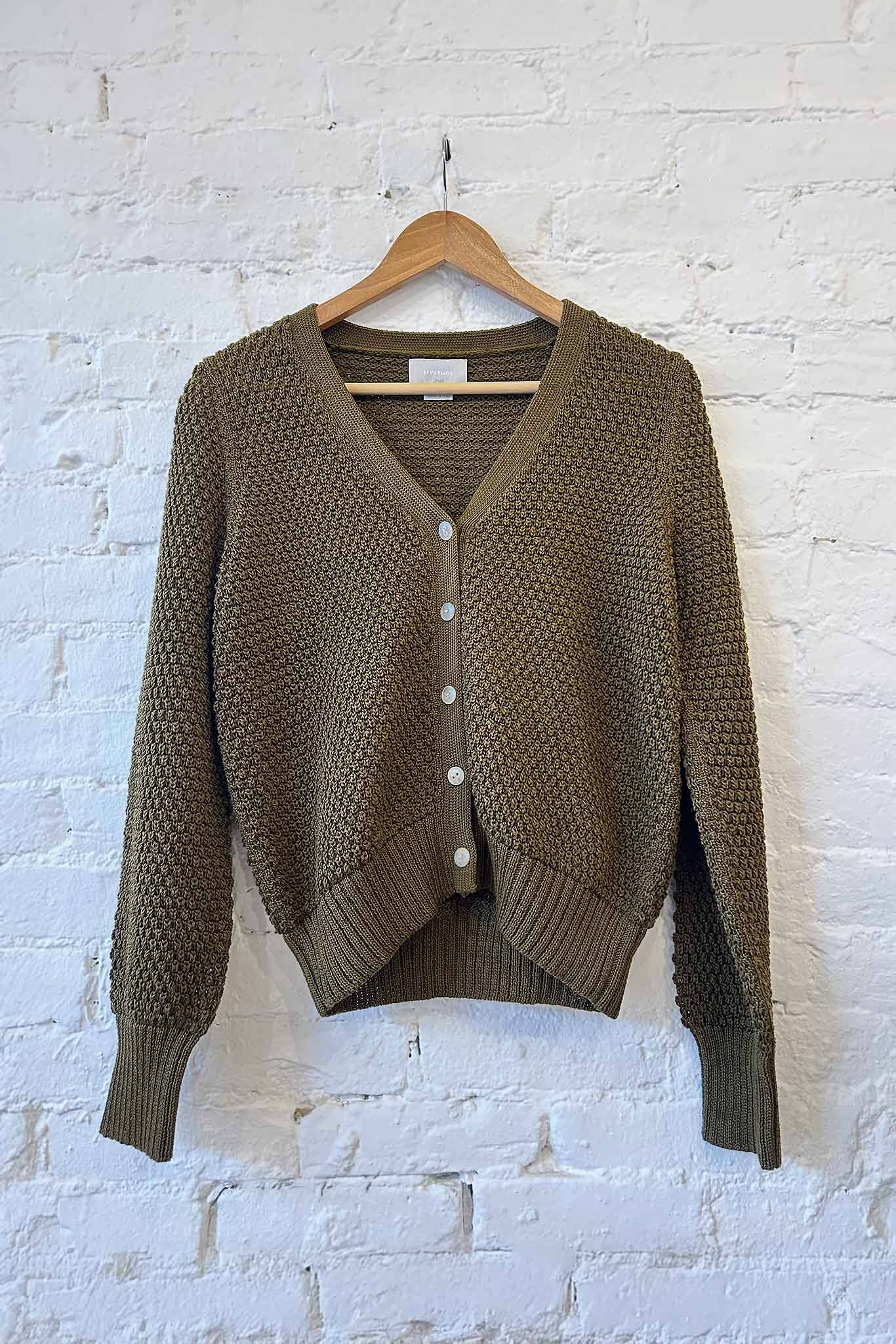 Olive green knitted cardigan on a wooden hanger against a white brick wall.