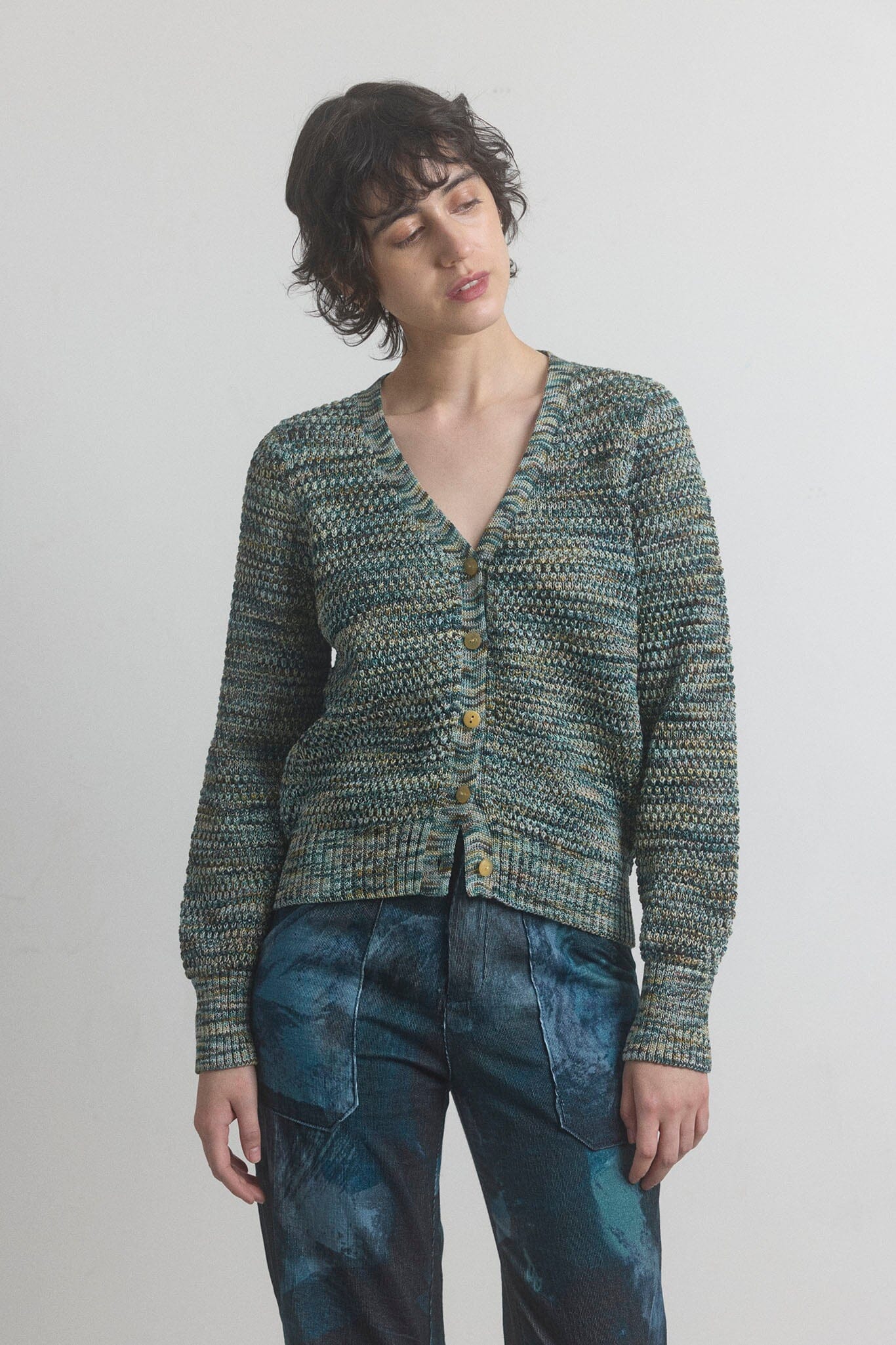 Woman wearing a green knitted cardigan and blue jeans against a plain background
