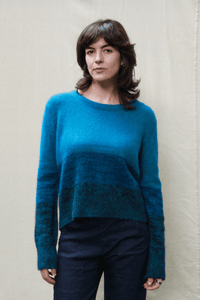 A woman wearing a teal and blue gradient knit sweater with dark navy pants, standing against a light beige fabric backdrop.