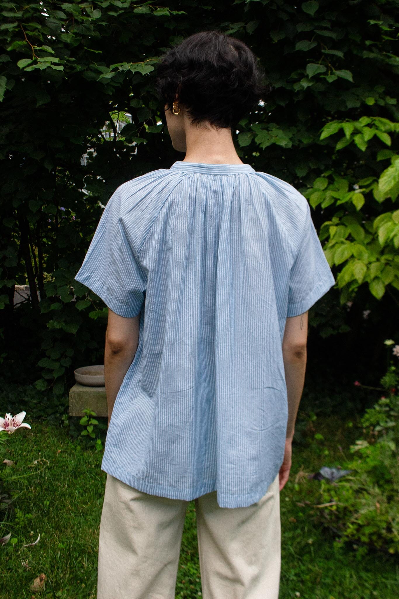 A person standing outdoors in a garden, viewed from the back, wearing a light blue short-sleeve top with a gathered, pleated back and beige pants.