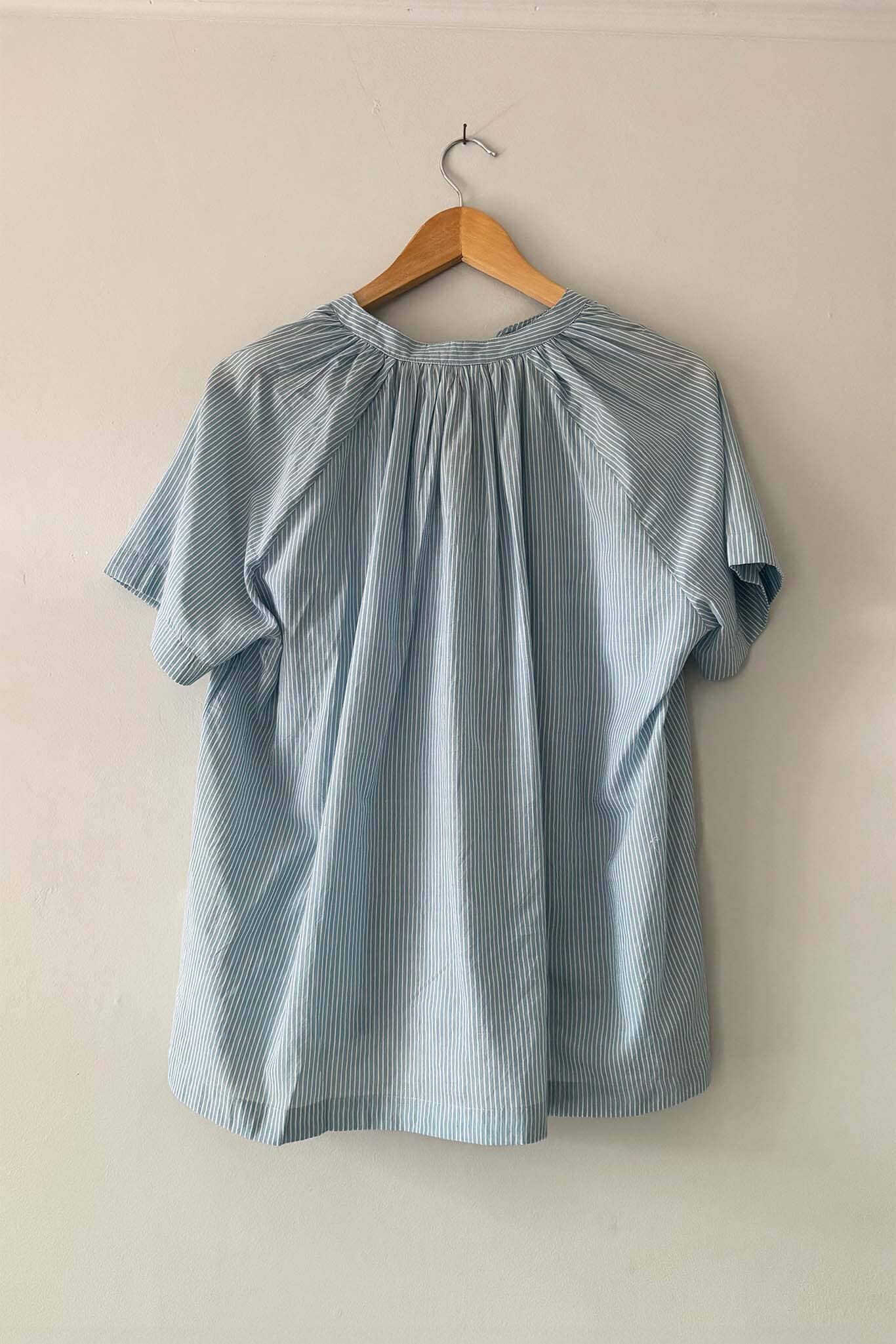 A light blue and white vertically striped short-sleeve blouse with a gathered, pleated neckline, hanging on a wooden hanger against a plain off-white wall.