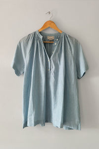 A light blue and white vertically striped short-sleeve blouse hanging on a wooden hanger against a plain light gray wall.