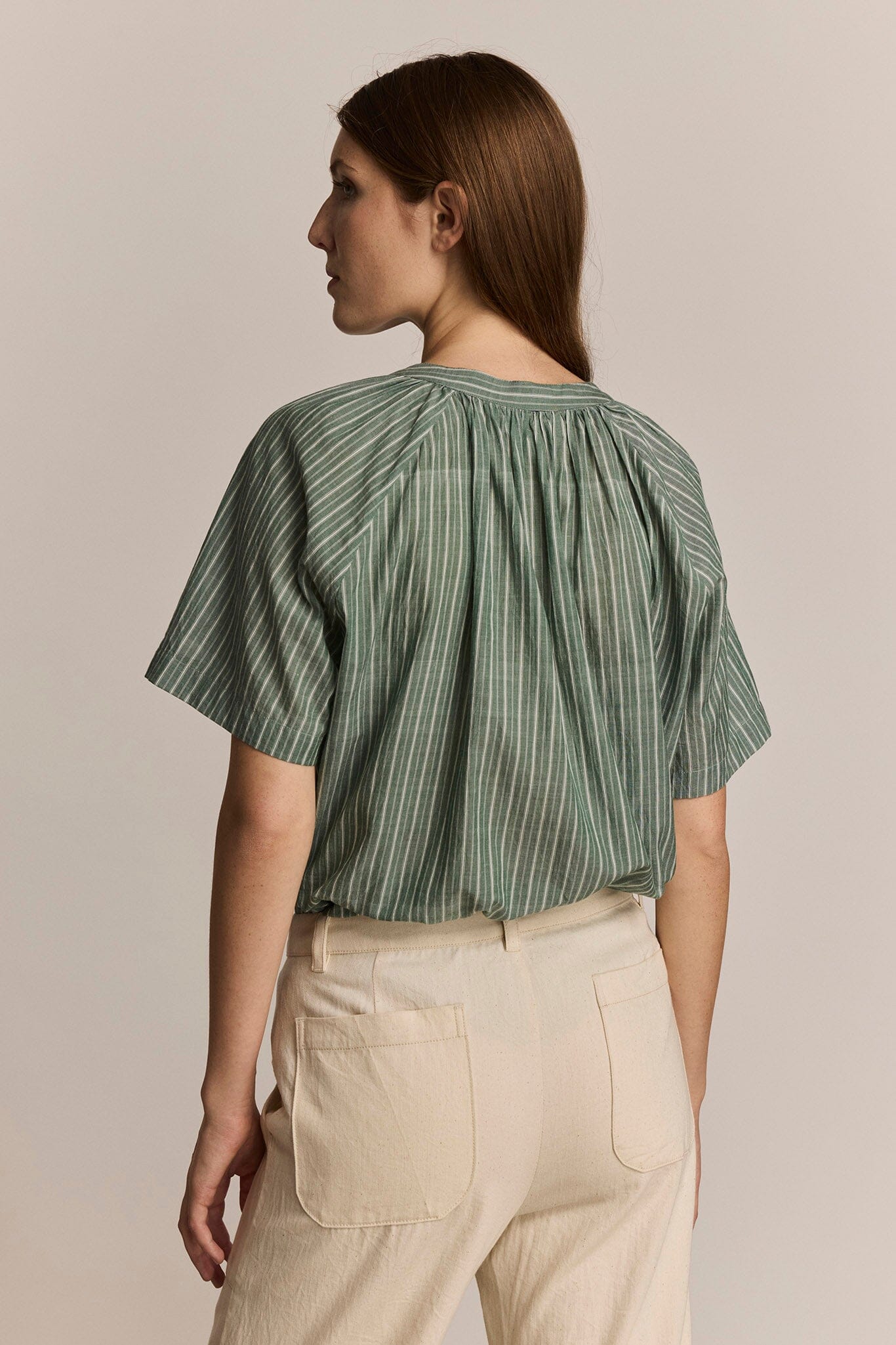 Woman facing the back wearing a green striped blouse and beige pants against a neutral background