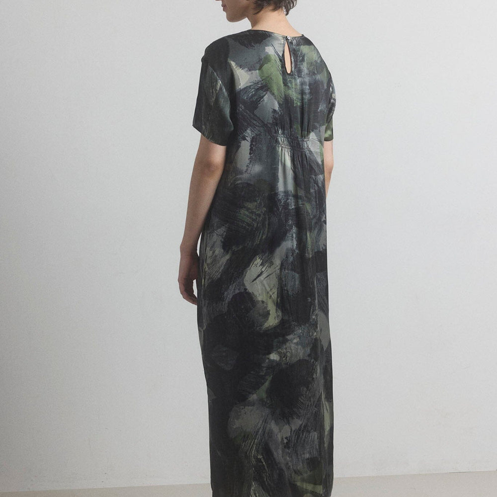 Woman facing the back wearing a long, dark green silk dress with a abstract pattern on a plain background