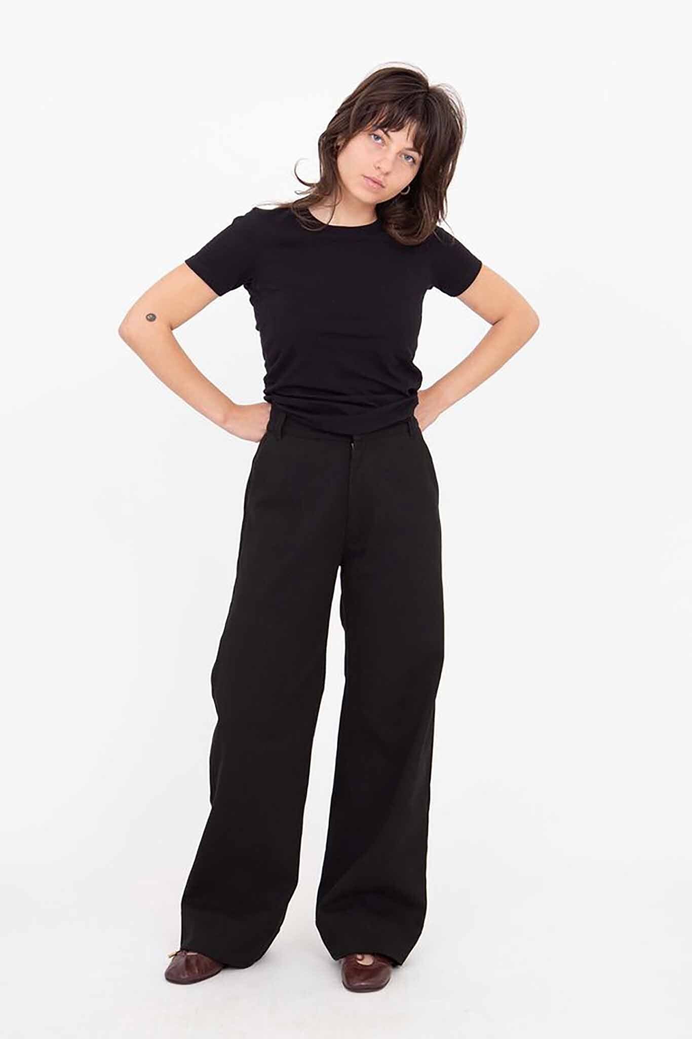 A woman standing, wearing black Riverside pants with a wide leg and a black top. She has short hair and is posing with her hands on her hips.