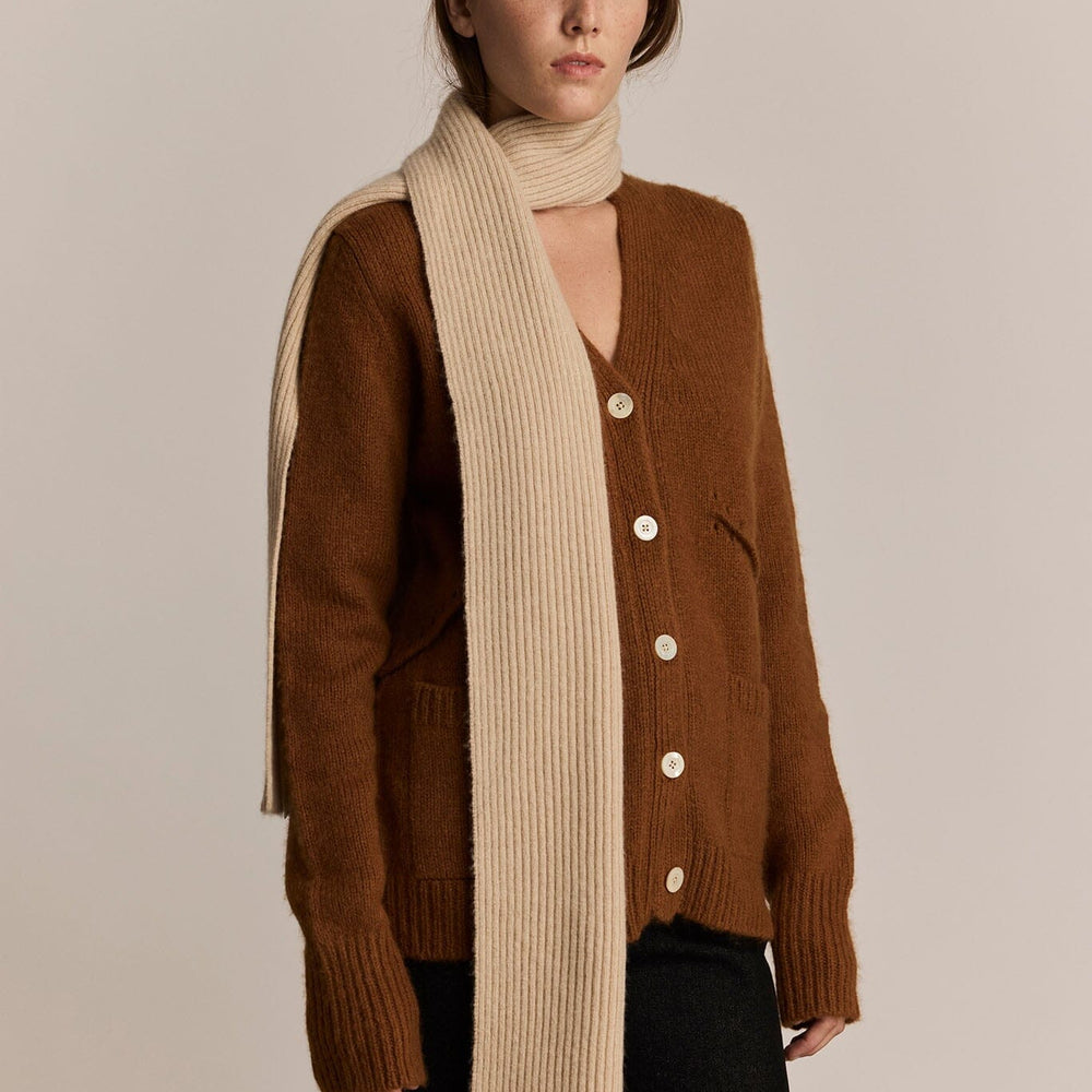 Woman wearing a brown cardigan and long beige knit scarf against a plain background