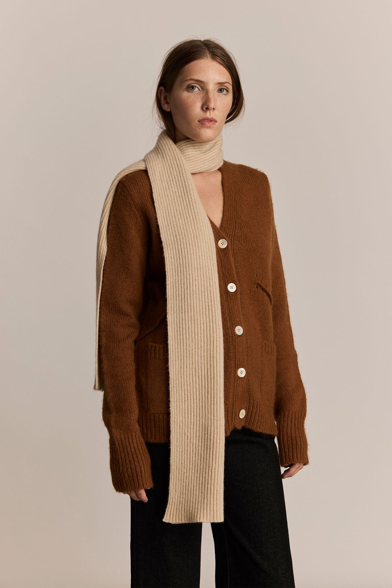 Woman wearing a brown cardigan and long beige knit scarf against a plain background