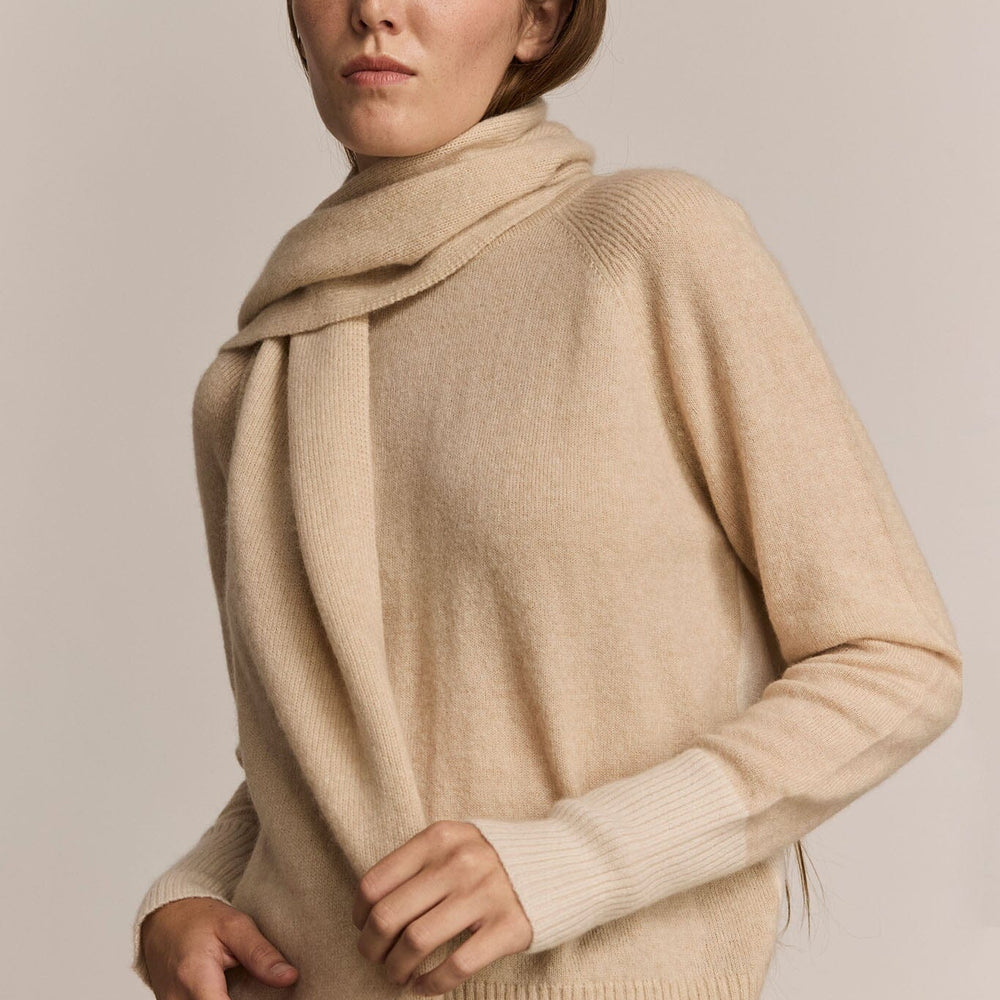 
                  
                    Woman wearing a long beige knit sweater and scarf against a plain background
                  
                