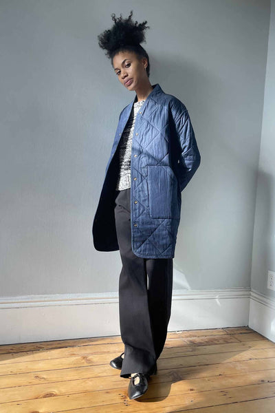 A woman wears the M.Patmos Sana quilted coat in a navy blue color. Long coat with snap buttons and two front pockets. Designed in Brooklyn, New York.