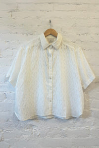 White textured short sleeve shirt on a wooden hanger against a white brick wall.