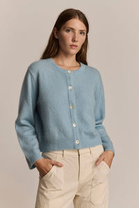 Woman wearing a light blue cardigan with mother of pearl buttons, beige pants against a plain background
